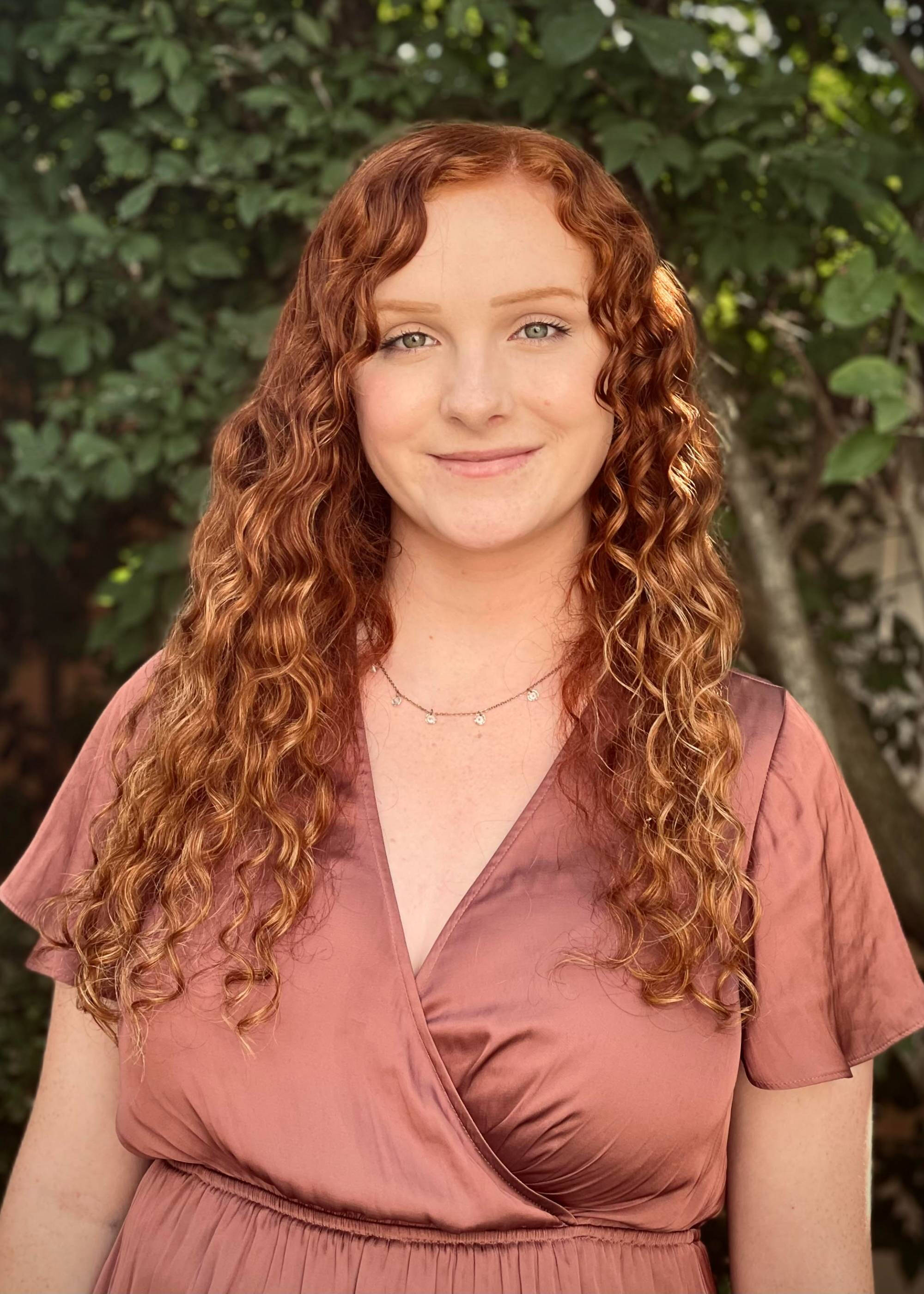 A student stands in front of a lush green background smiling.